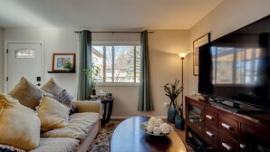 A living room with a sofa, a coffee table, a TV, and a window with curtains. - Loveland Home w/ Private Hot Tub + Wood Fireplace! (Loveland)