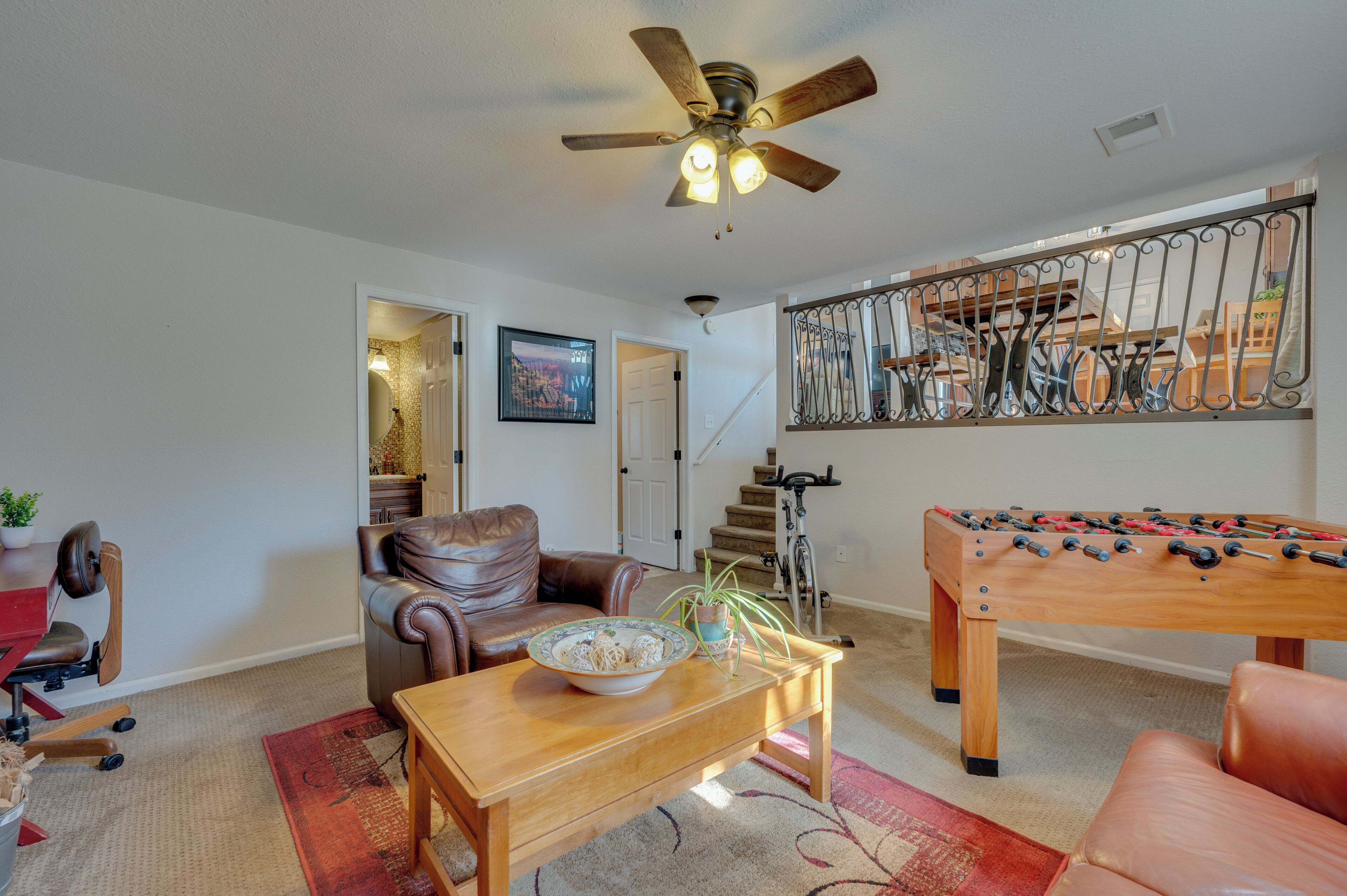 A living room with a wooden coffee table, a brown leather sofa, a foosball table, a ceiling fan, and a staircase leading to another room.
