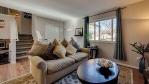 A living room with a beige sofa, a coffee table, and a staircase leading to an upper level. - Loveland Home w/ Private Hot Tub + Wood Fireplace! (Loveland)