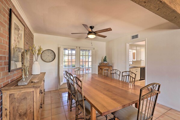 A dining room with a wooden table, chairs, a ceiling fan, and a brick wall with a framed picture.