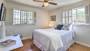 Une chambre à coucher avec un lit, un ventilateur de plafond, une fenêtre avec des volets, une table de chevet avec une lampe et un tableau encadré accroché au mur.