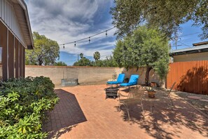 A patio area with brick flooring, two blue lounge chairs, a fire pit, and string lights.