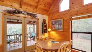 A wooden cabin interior with a dining table, chairs, a rocking chair, a window, and a door leading to an outdoor deck.