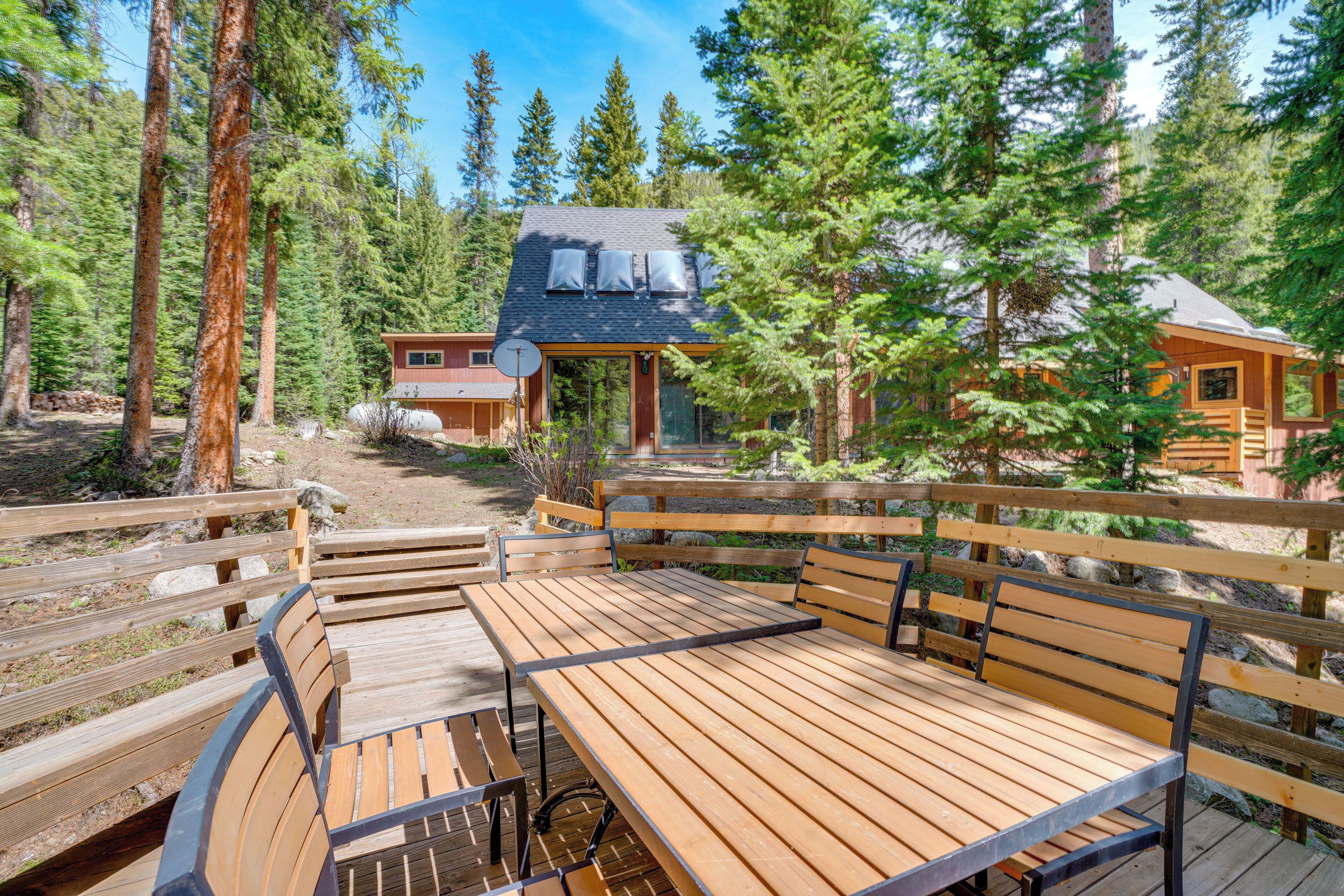 A wooden deck with benches and a table, surrounded by tall trees and a cabin with a satellite dish.
