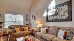 A living room with two brown sofas, a coffee table with decorative items, a large wall art piece, and a window with blinds. - Enchanted Forest Cottage w/ Hot Tub! (Hiawassee)