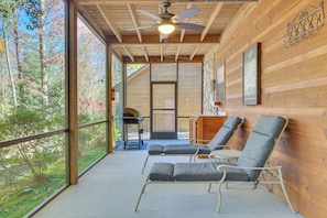 A wooden cabin with a balcony, two lounge chairs, a ceiling fan, and a view of the forest.