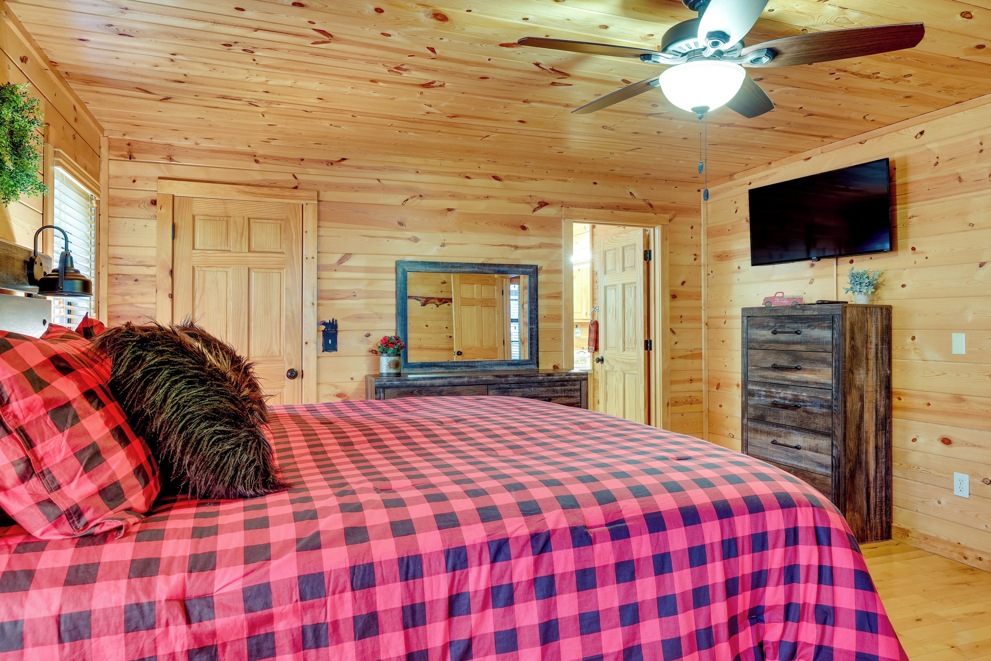 A bedroom with a checkered bedspread, a ceiling fan, a television, and wooden walls.