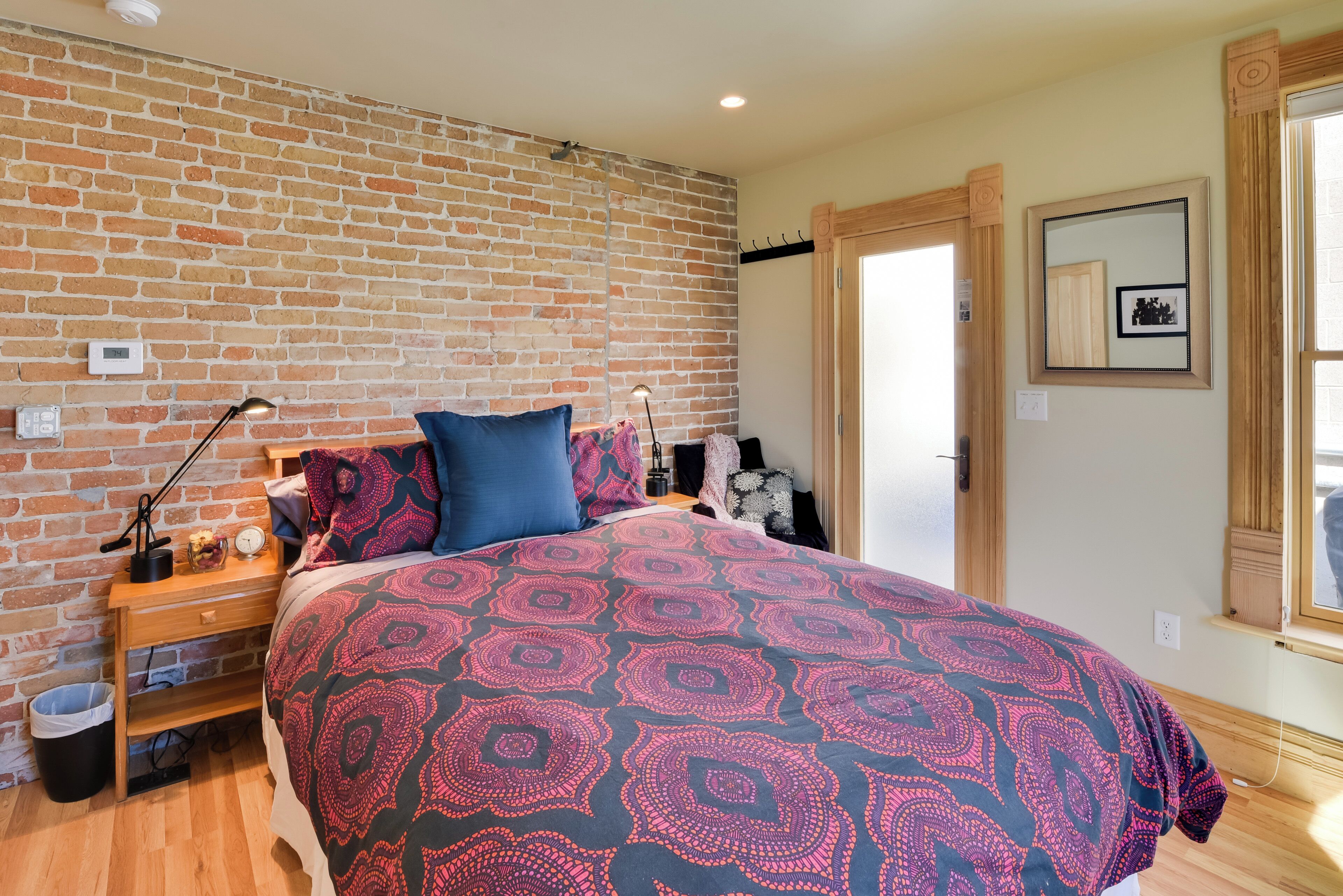 A bedroom with a brick wall, a bed with a patterned bedspread, a nightstand with a lamp, and a wooden dresser.