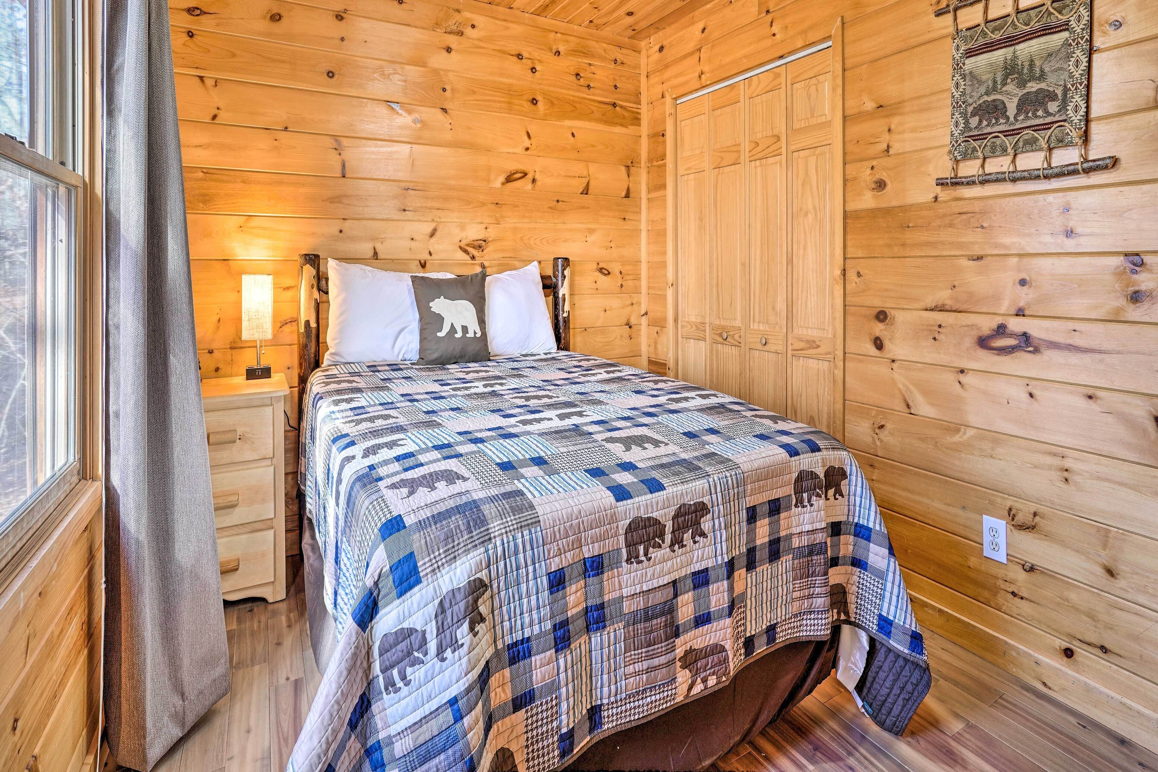 A wooden cabin bedroom with a bed, a nightstand, a window with curtains, and a wall-mounted tapestry.