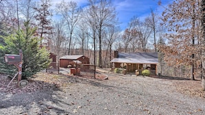 A rustic cabin with a metal roof, surrounded by trees and a gravel driveway. - 'serenity Cabin' w/ Furnished Deck (Mineral Bluff)