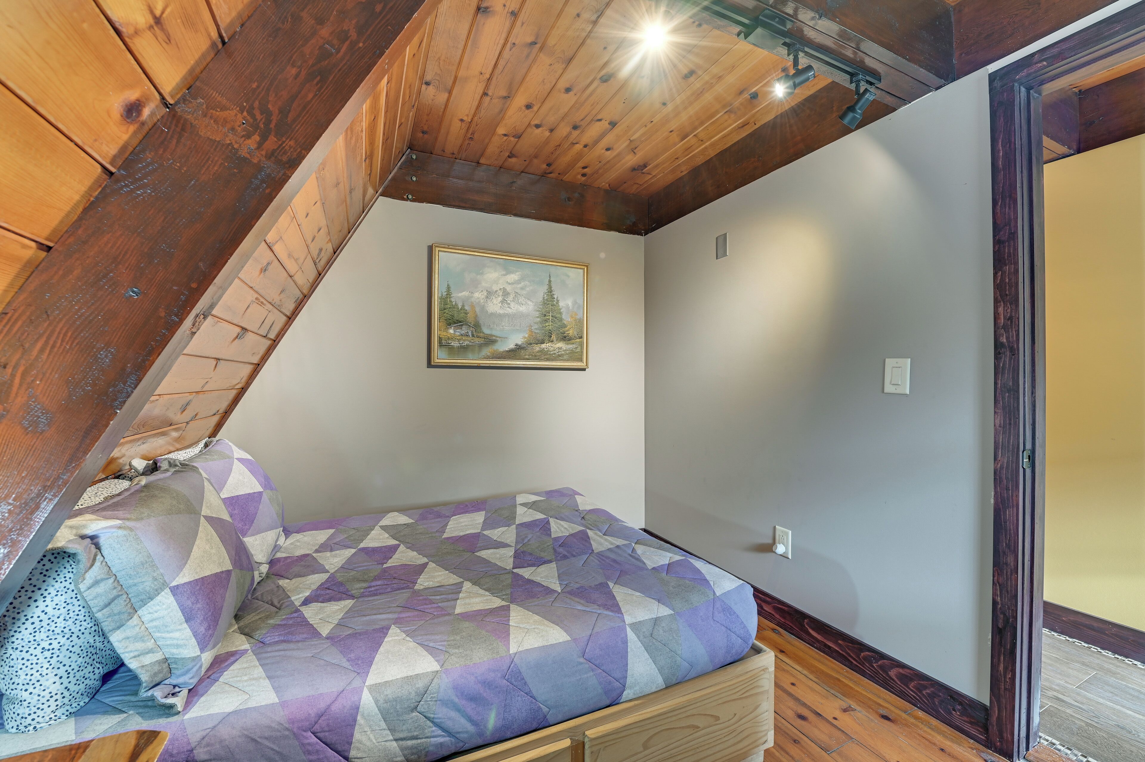 A bedroom with a wooden ceiling, a bed with a patterned bedspread, a painting on the wall, and a wooden floor.