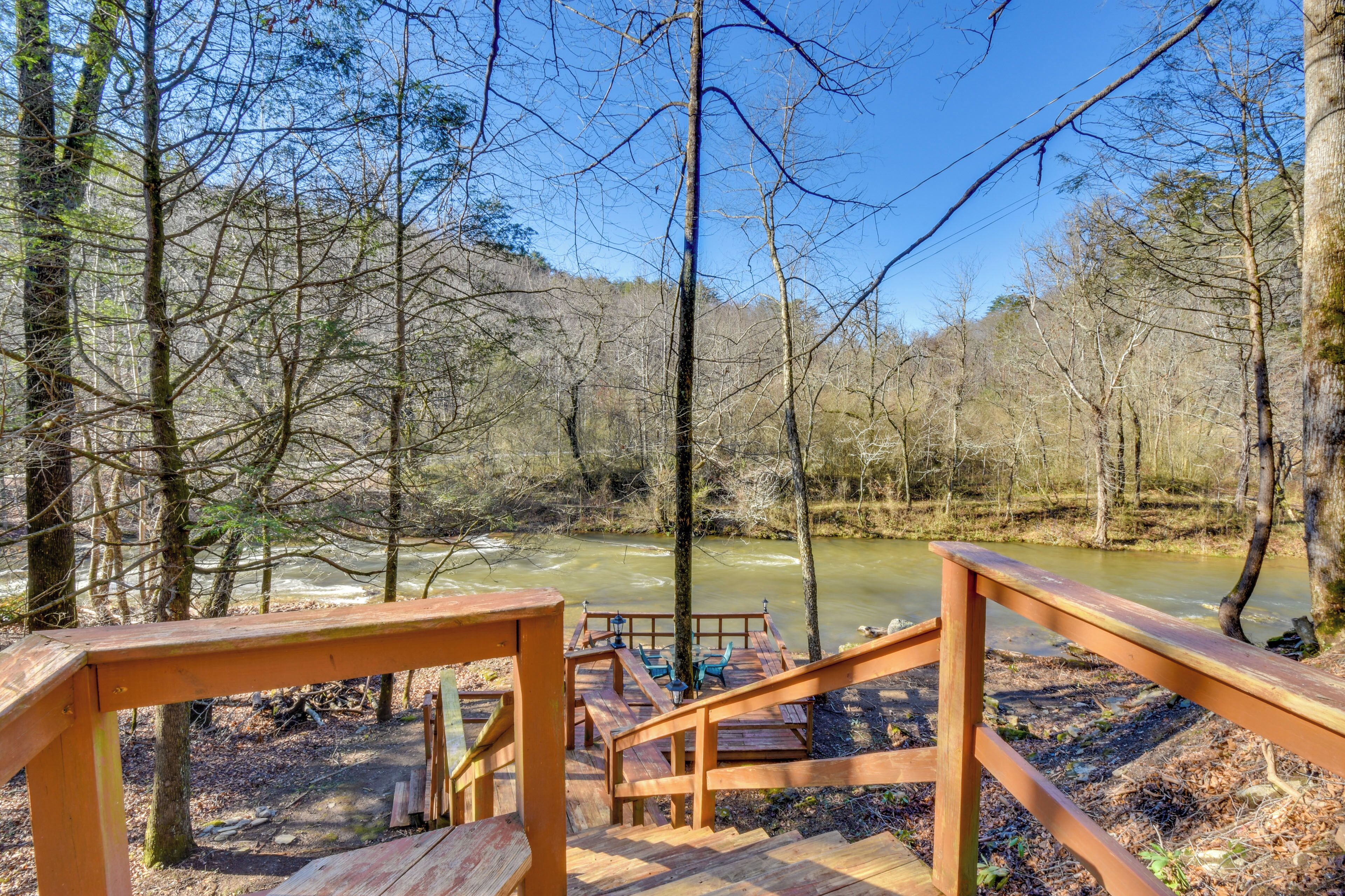 A wooden deck with steps leading to a river view, surrounded by trees.