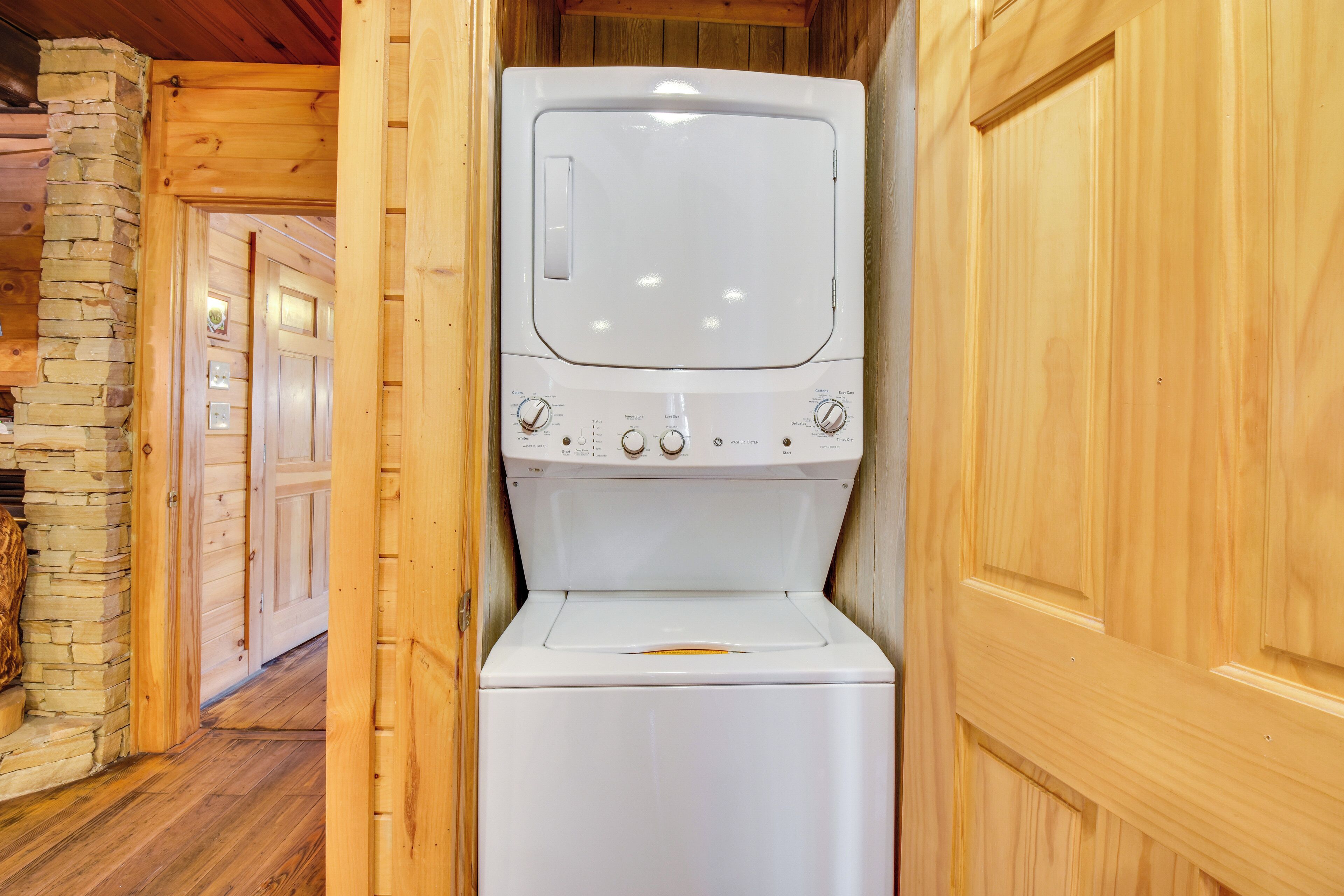 A compact, white washing machine in a wooden cabin with a stone wall and a wooden door.