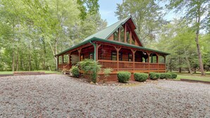 Une cabane en bois avec un toit vert, entourée d’une allée en gravier et d’une zone boisée.