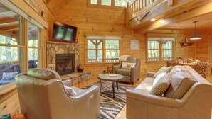 Un intérieur de chalet chaleureux avec une cheminée en pierre, des canapés en cuir, un coin repas et un téléviseur fixé au mur.