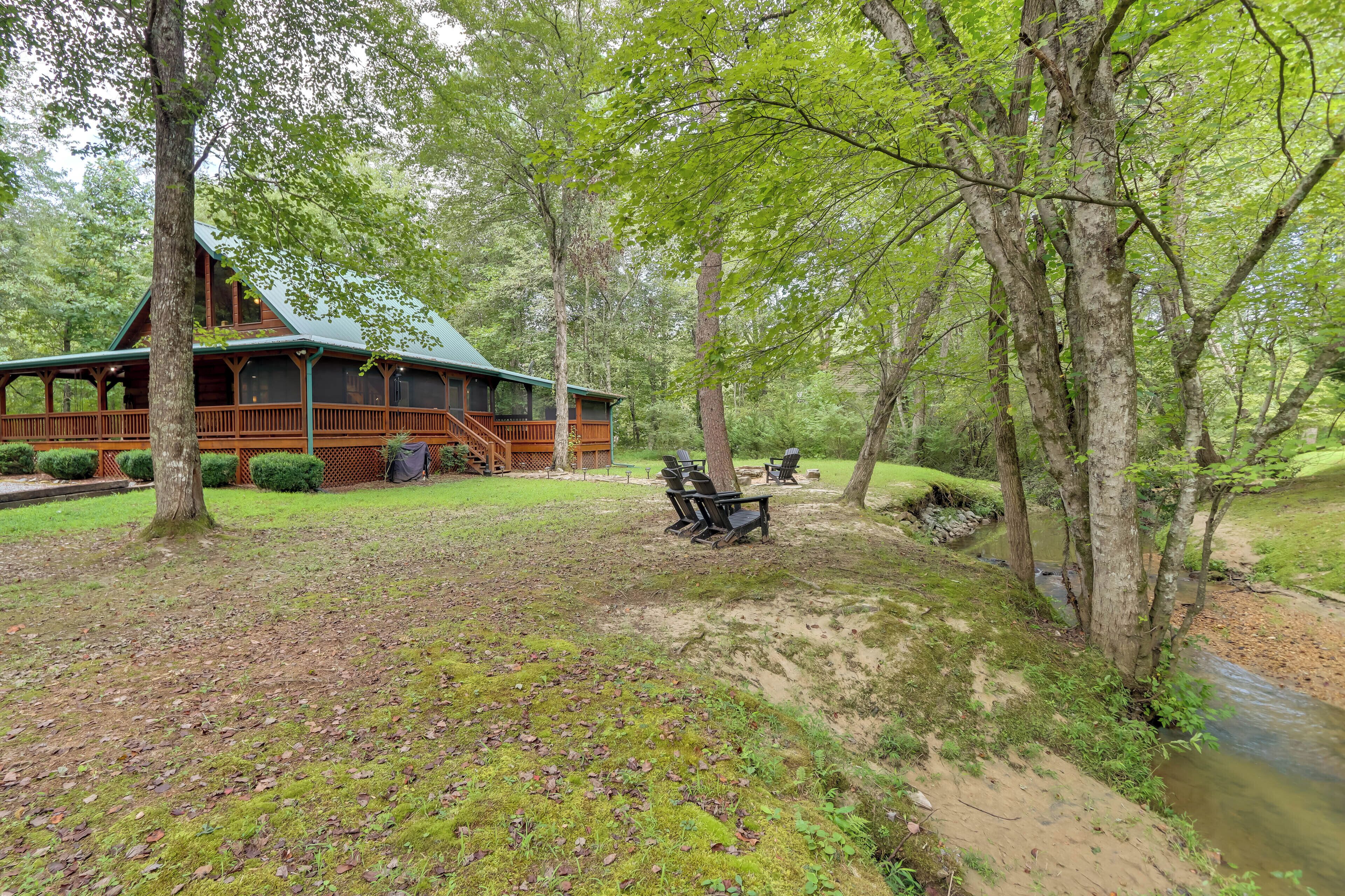 A cabin with a porch and outdoor seating area, surrounded by trees and a stream.