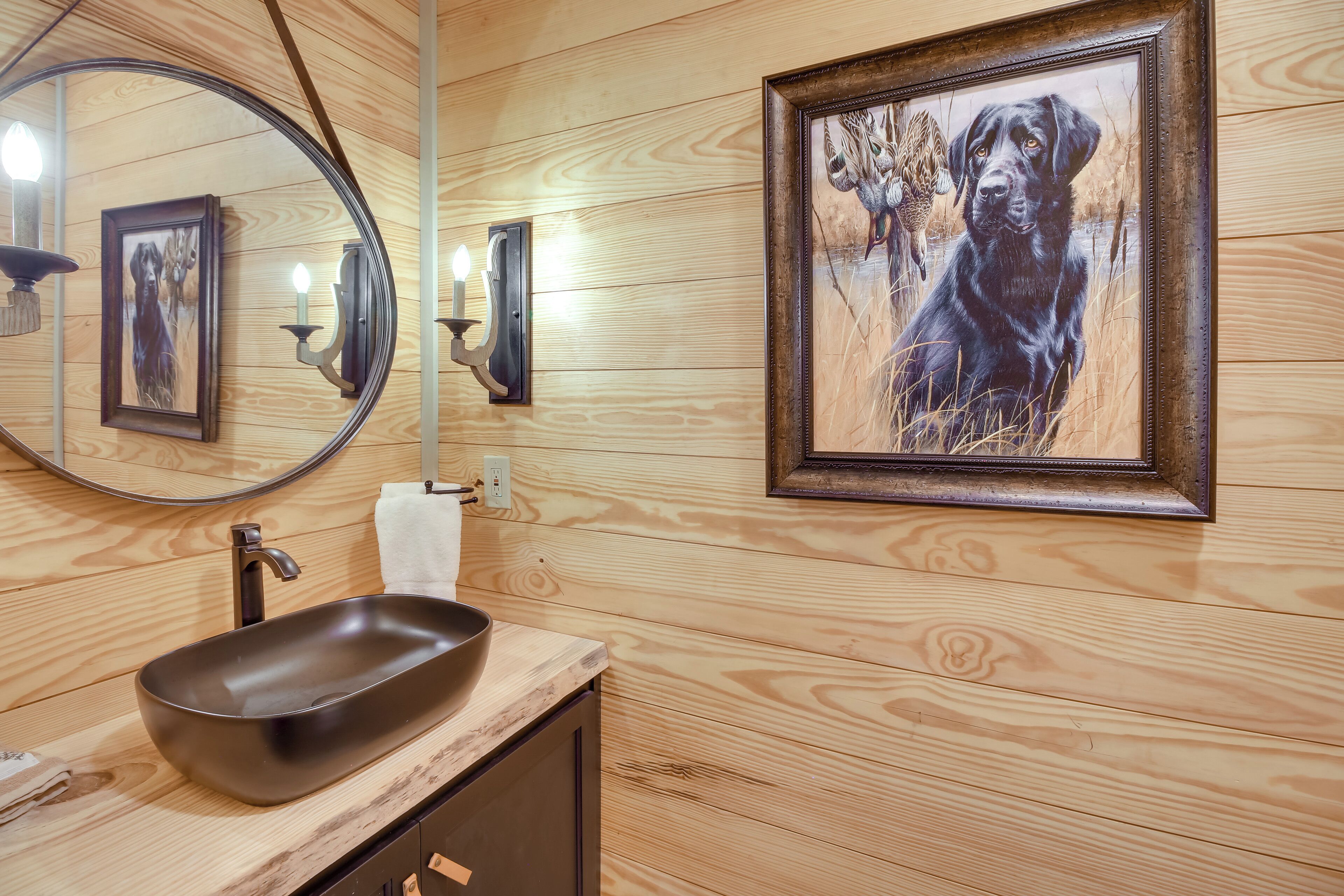 A wooden-paneled bathroom with a round mirror, a black sink, and a framed painting of a dog.