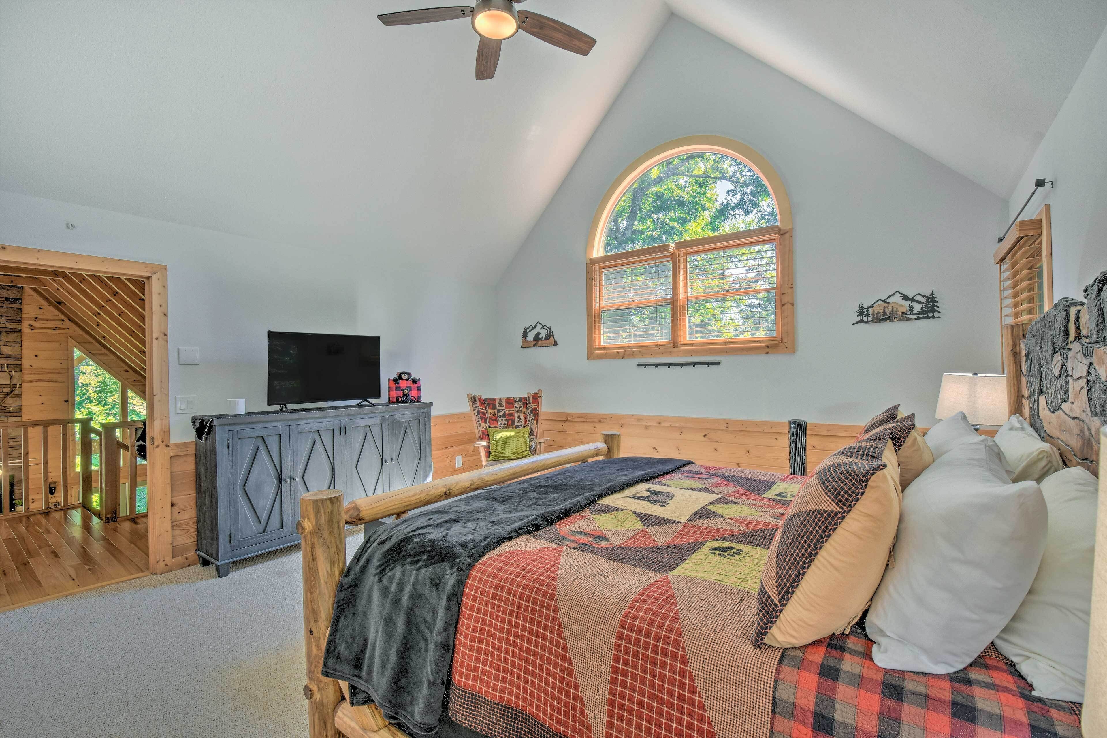 A bedroom with a large bed, a ceiling fan, a TV on a cabinet, and a window with a view of trees.