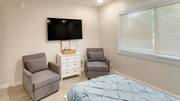 A bedroom with a bed, two armchairs, a TV, and a window with blinds. - Bright Jekyll Island Home w/ Deck < 1 Mi to Beach (Jekyll Island)
