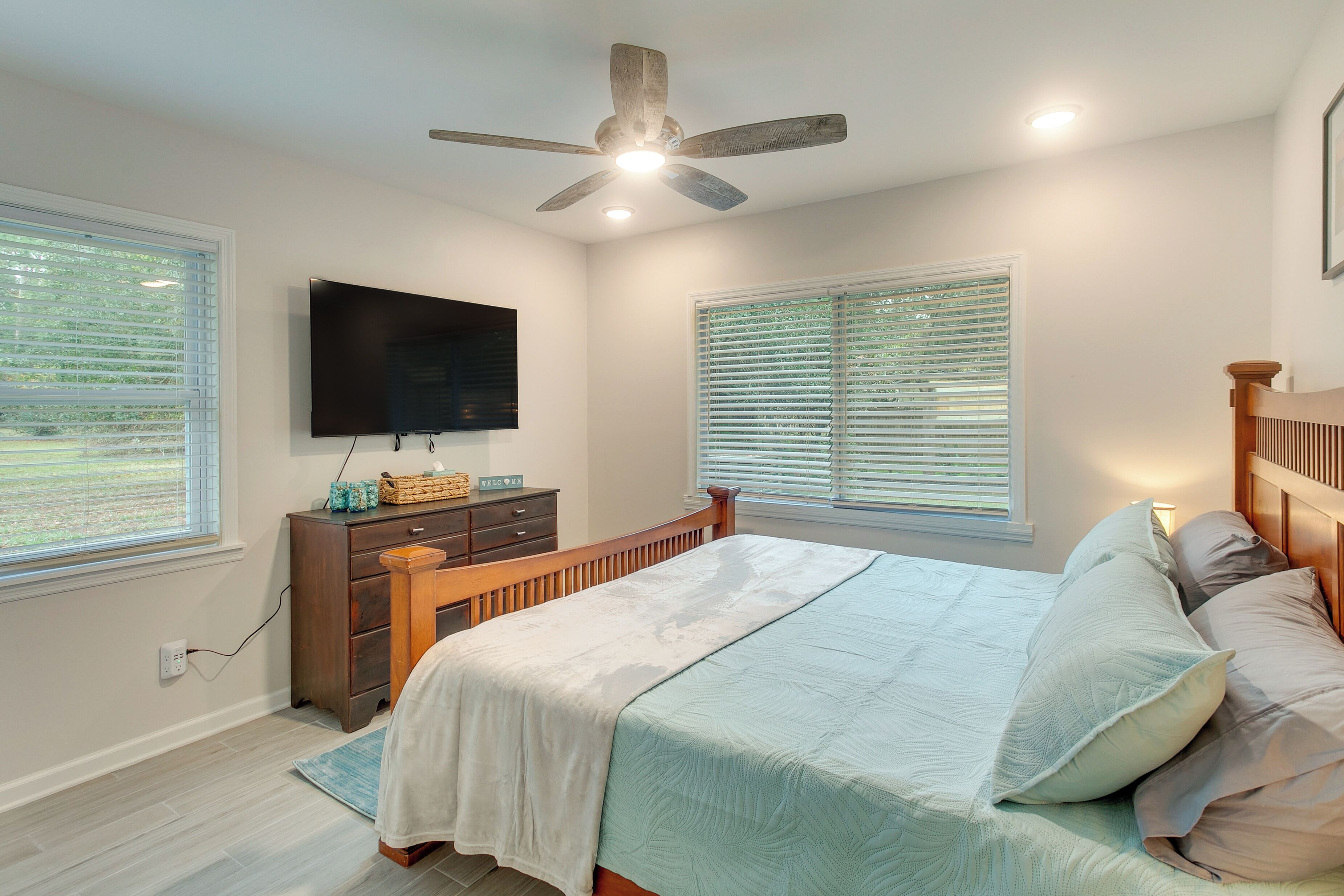 Une chambre à coucher avec un lit, un ventilateur de plafond, un téléviseur fixé au mur et des fenêtres équipées de stores.