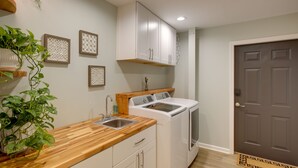 Une cuisine compacte avec des meubles blancs, un plan de travail en bois, un lave-linge et un sèche-linge intégrés, et une plante en pot.