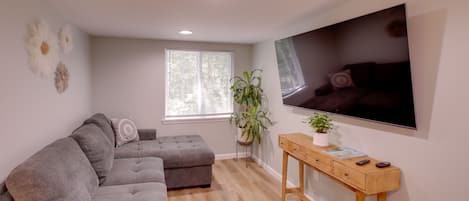 A modern living room with a grey sectional sofa, a wooden console table, a large flat-screen TV, and a window with blinds.