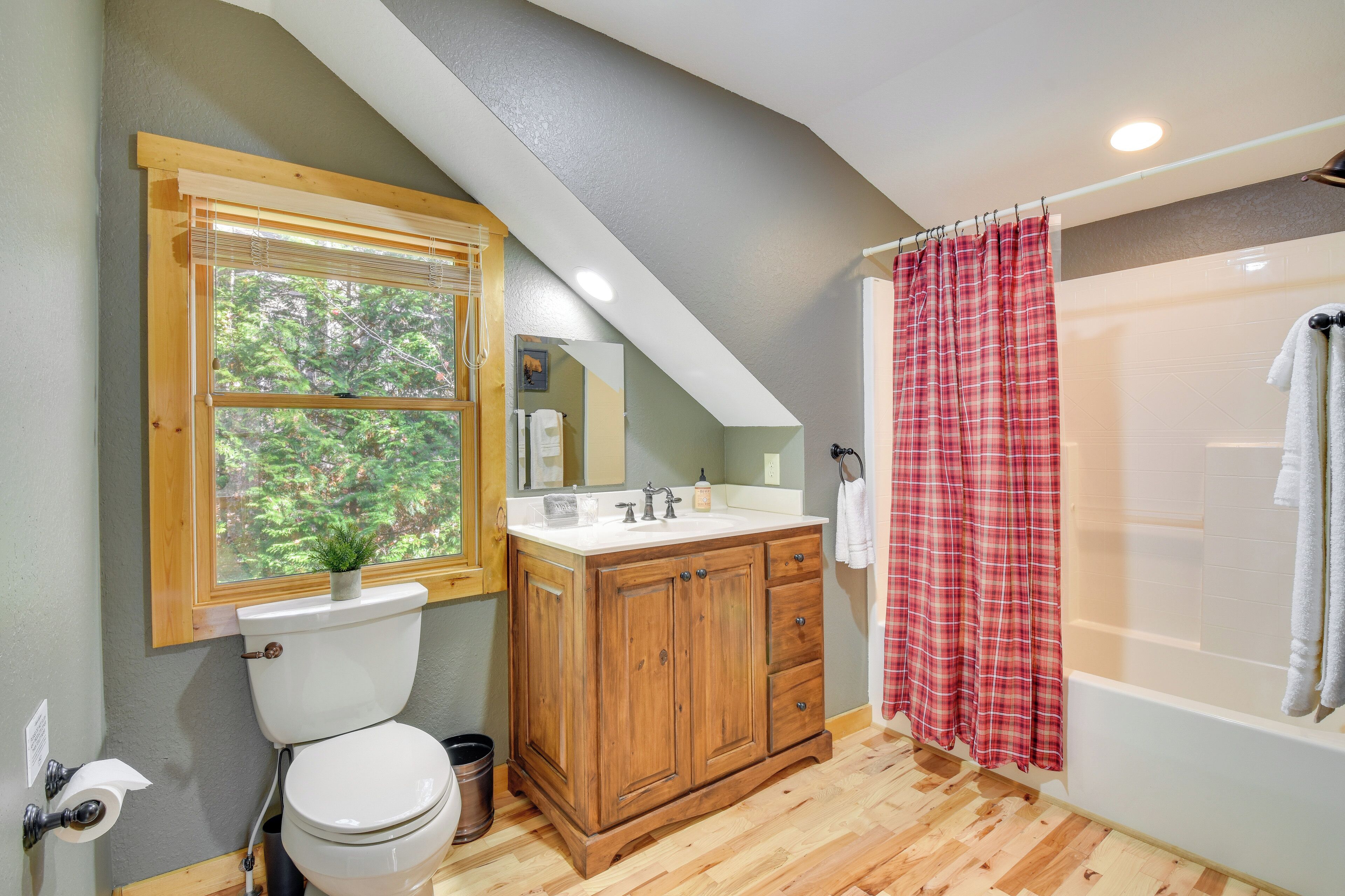 A bathroom with a wooden vanity, a toilet, a window with a view of trees, and a red plaid shower curtain.