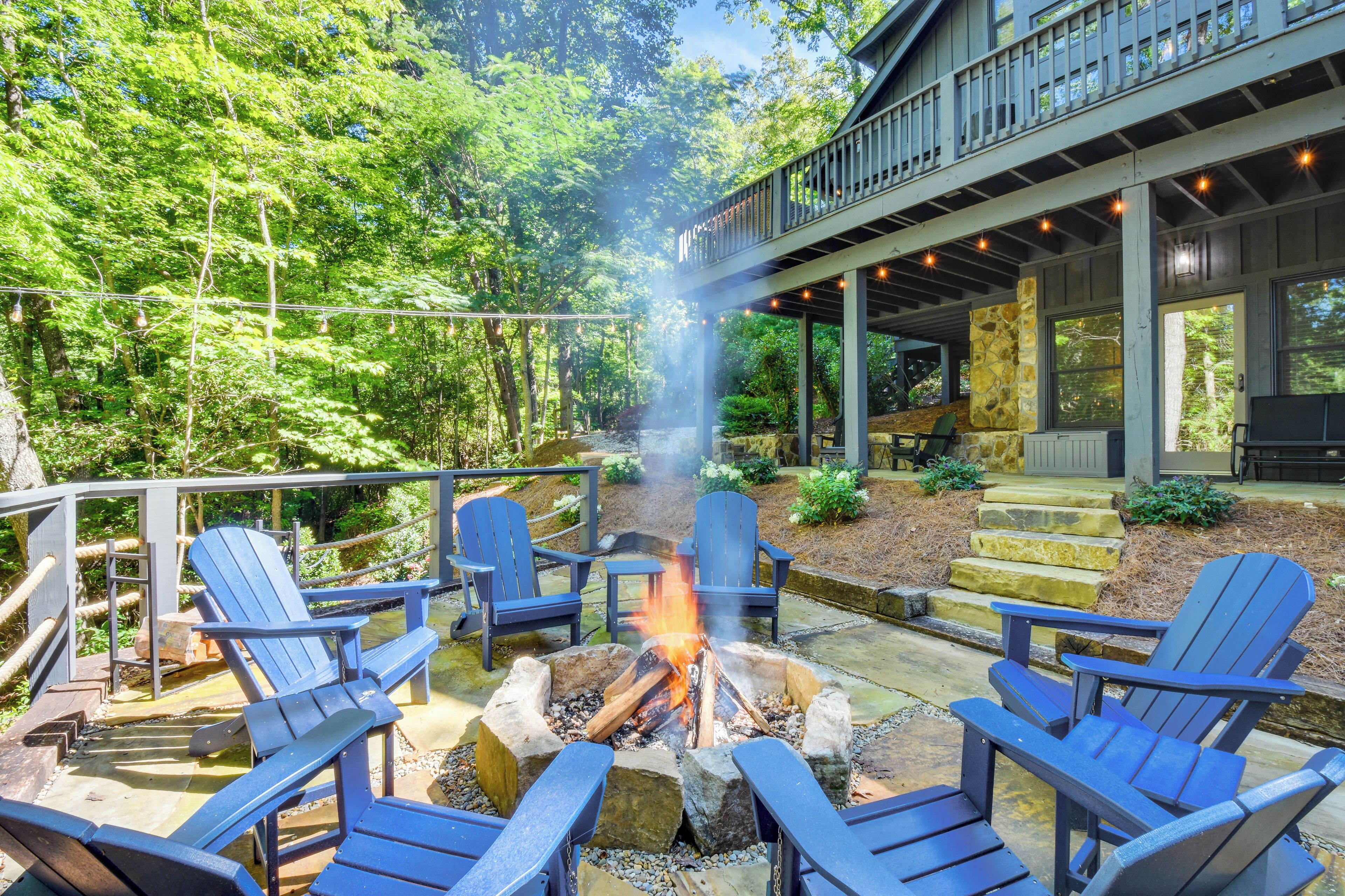 A fire pit surrounded by Adirondack chairs, a stone staircase, and a wooden deck with string lights.