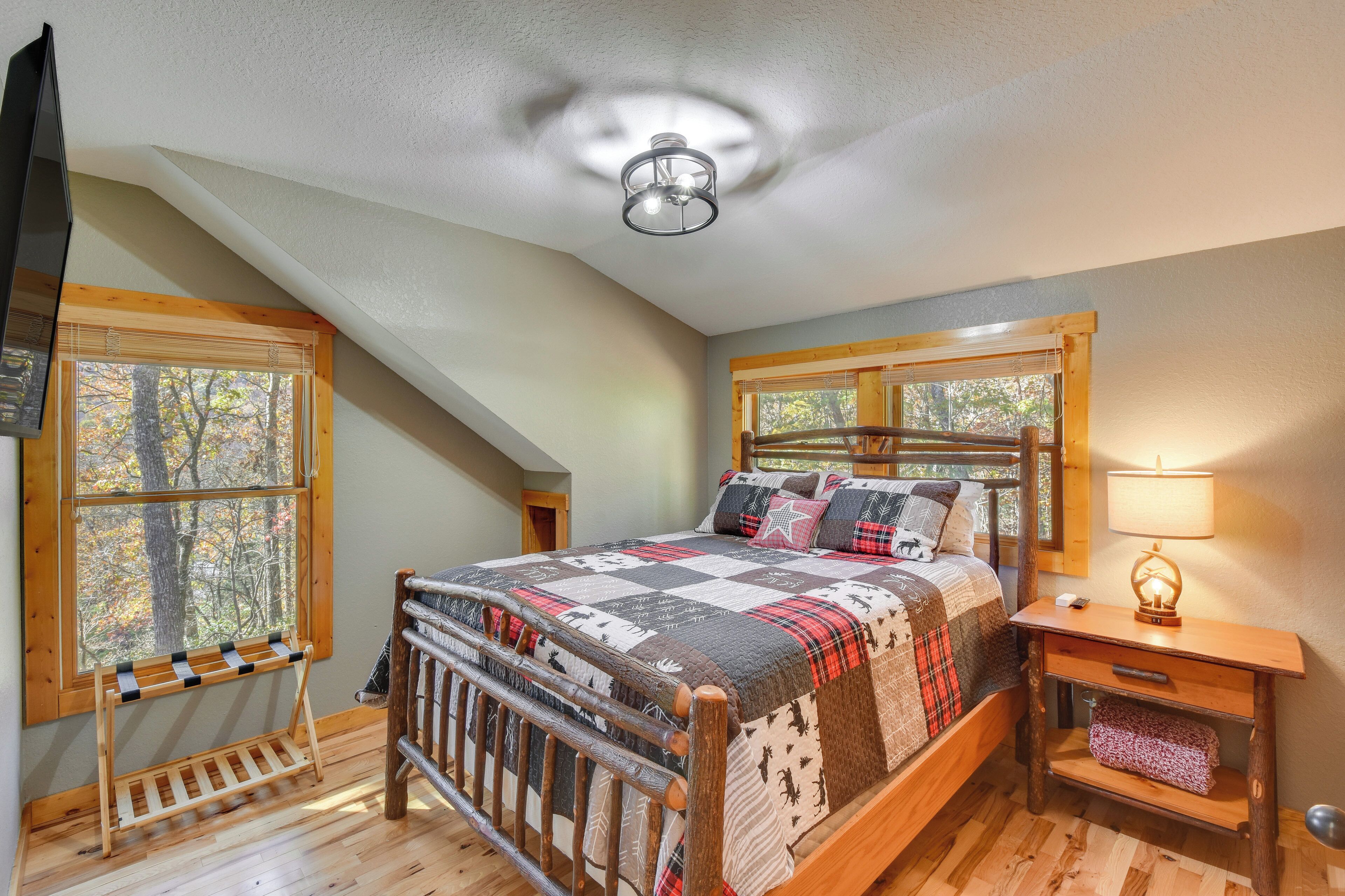 A bedroom with a wooden bed, a nightstand with a lamp, a window with a view of trees, and a ceiling fan.