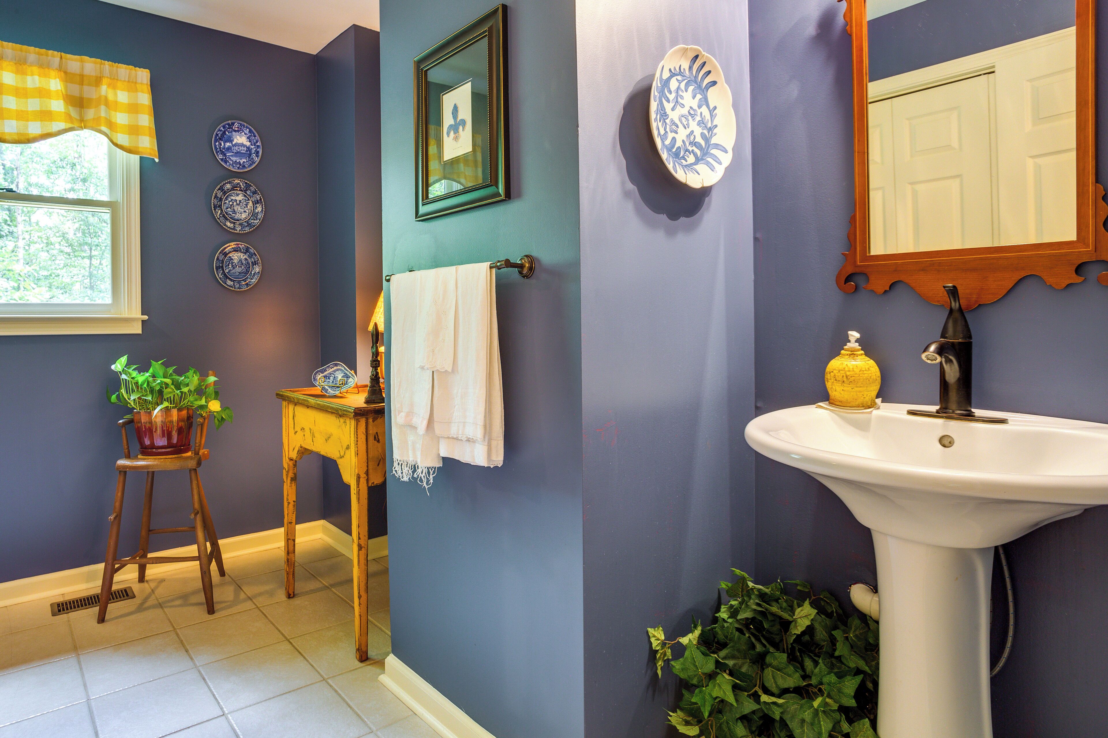 Une salle de bain avec un lavabo, un miroir et un porte-serviettes. Les murs sont décorés d’assiettes et il y a une fenêtre avec un rideau à carreaux.