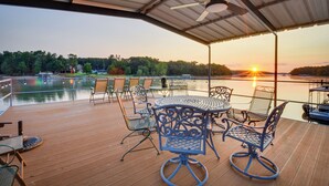 Una terraza techada al aire libre con una mesa y sillas, con vista a un lago con barcos y árboles al fondo.