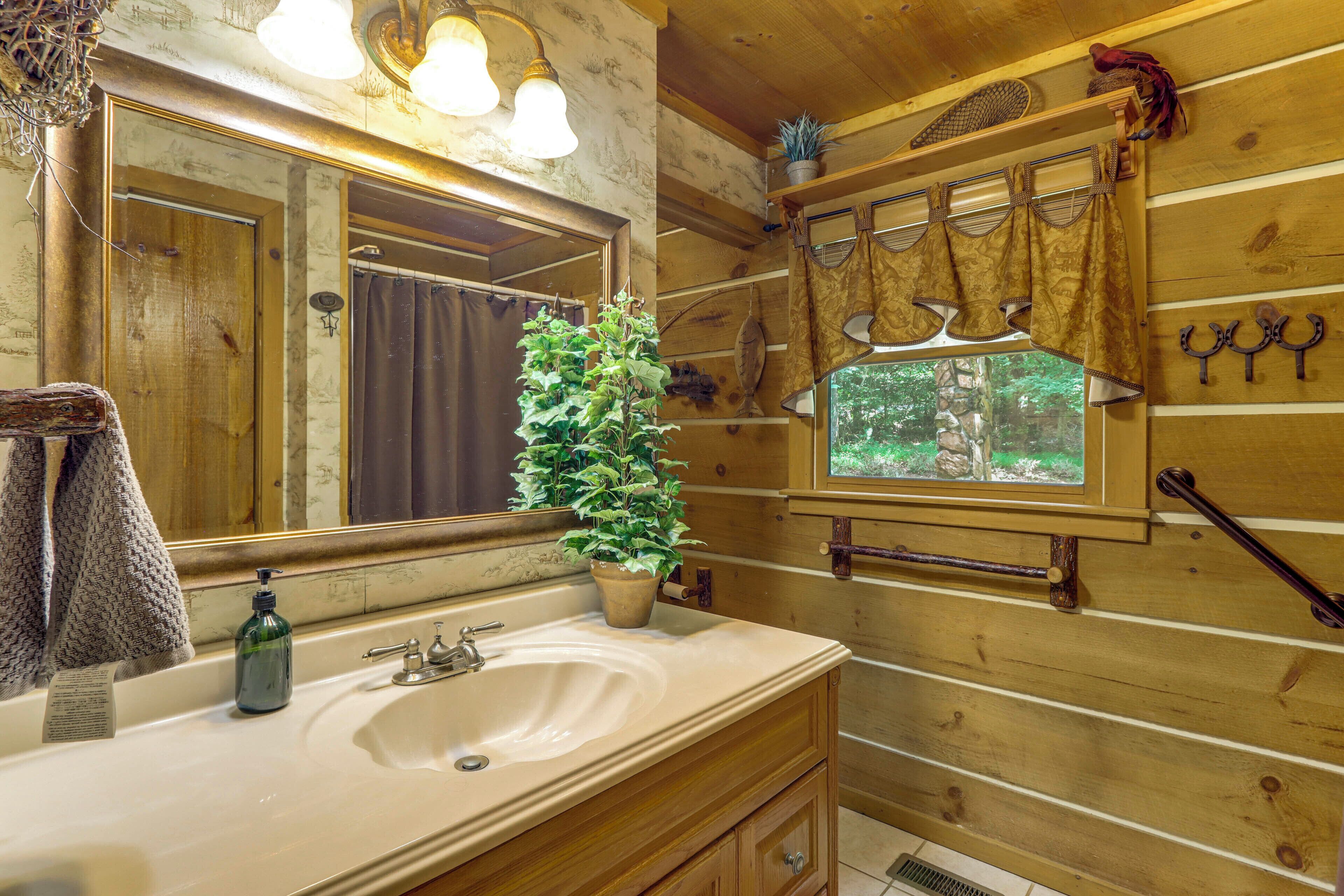 A wooden cabin bathroom with a sink, mirror, towel rack, potted plant, and window with curtains.