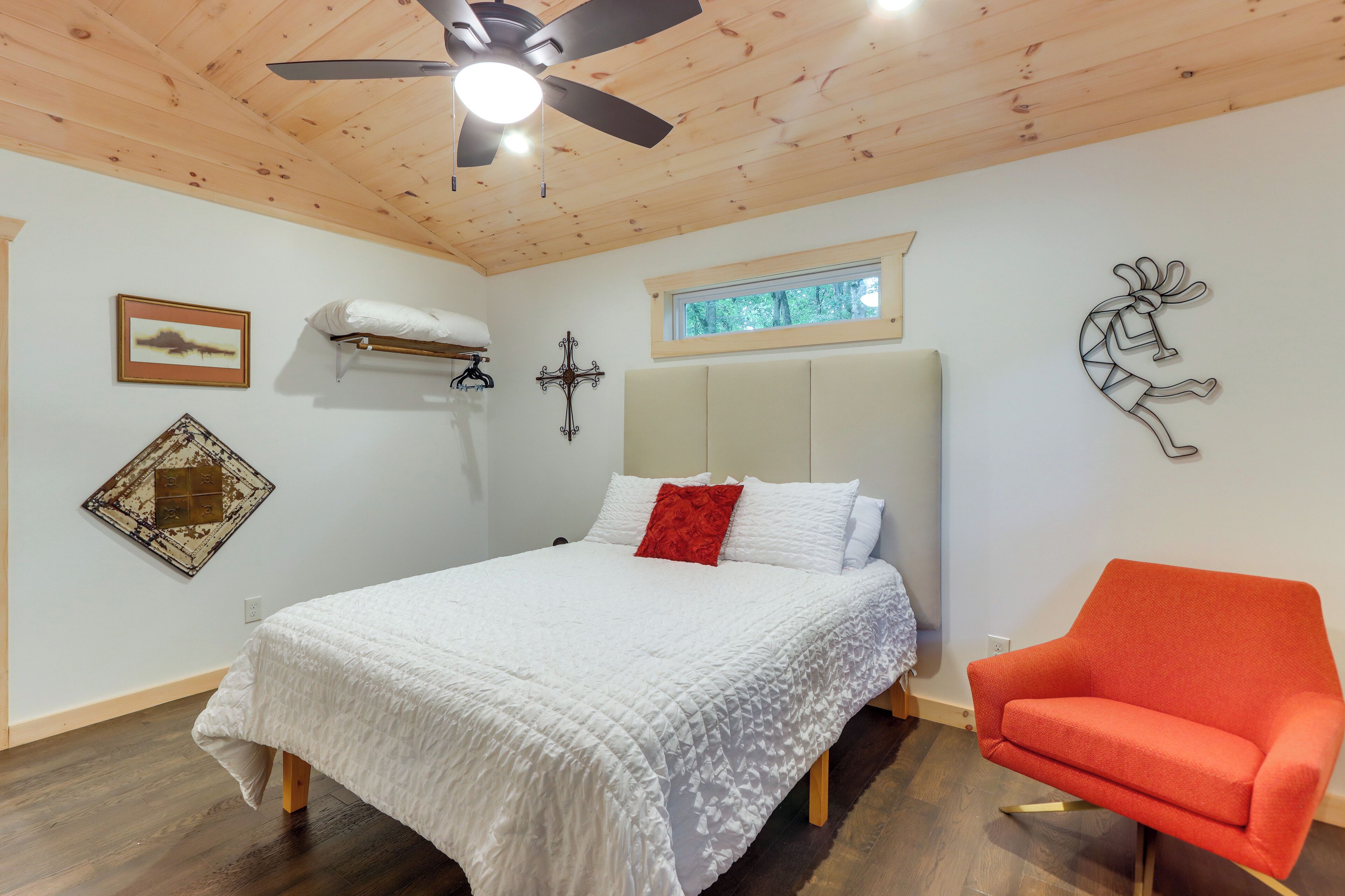 A bedroom with a bed, a red chair, a ceiling fan, a window, and wall decorations.