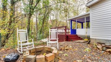 A rustic outdoor seating area with two white chairs and a brick fire pit, surrounded by a natural forest setting.