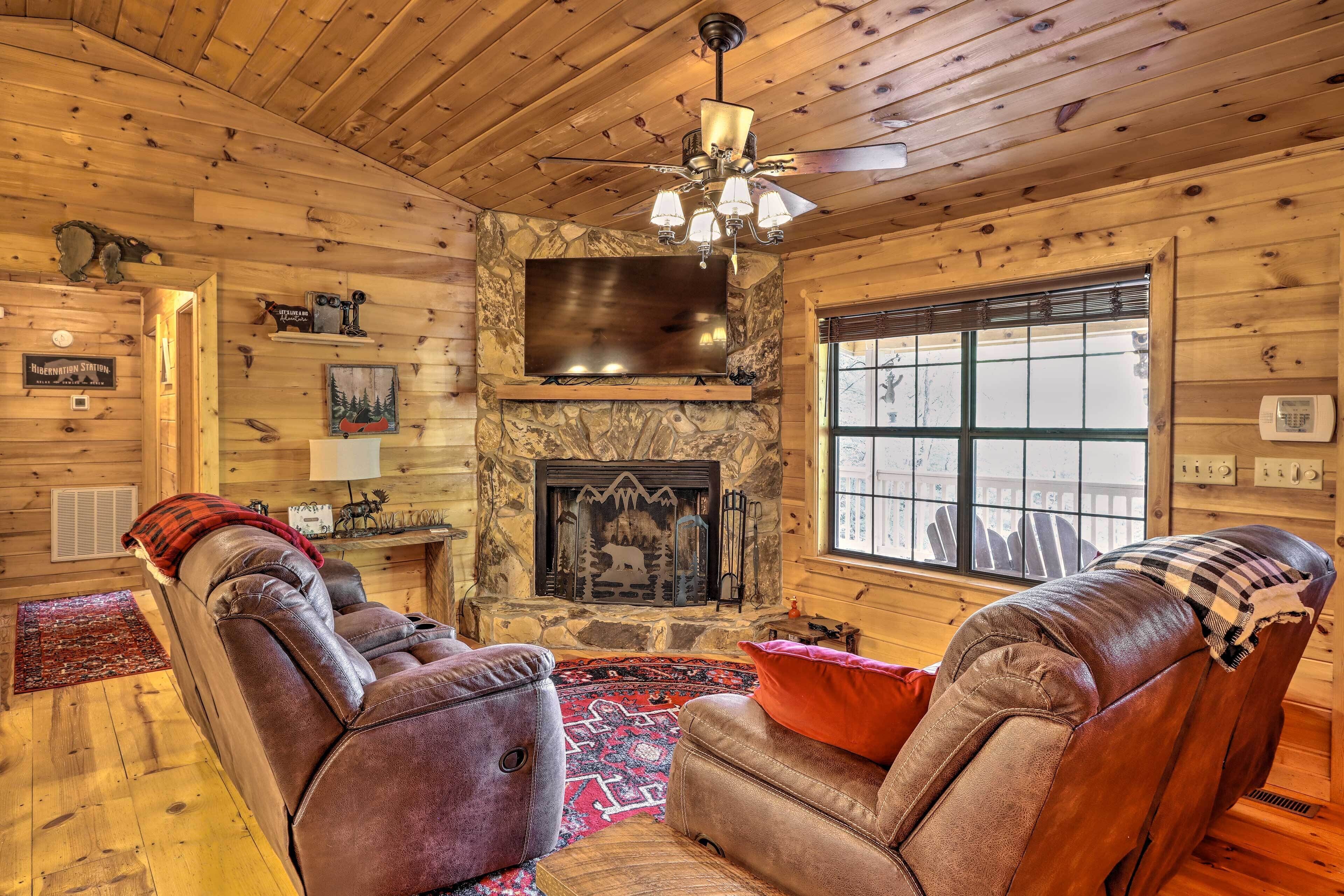 A log cabin interior with a fireplace, leather recliners, a ceiling fan, and a stone wall.