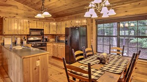 Une cuisine rustique aménagée dans une cabane en bois, avec un îlot central, une table à manger entourée de chaises et une grande fenêtre donnant sur la forêt.