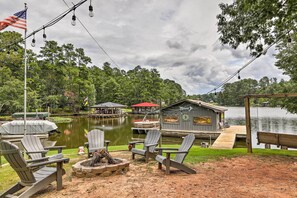 Una terraza frente al lago con sillas Adirondack, un hogar para fogata y un muelle que se adentra en el agua.