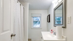 Ein Badezimmer mit weißer Tür, einer Duschvorhang mit geometrischem Muster, einem Fenster mit Jalousien, einem Waschbecken mit Spiegel und Toilettenartikeln auf der Ablagefläche.