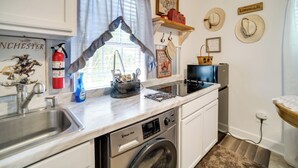 Eine kleine Küche mit Spüle, Herd und Geschirrspüler. Ein Feuerlöscher, ein Fenster mit Vorhängen sowie ein Regal mit Hüten und einem Korb sind vorhanden.