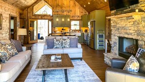 A spacious living area with a high ceiling, wooden walls, a stone fireplace, and a kitchen with green cabinets. - Riverside Mtn Retreat w/ Hot Tub & Game Room! (Blue Ridge)
