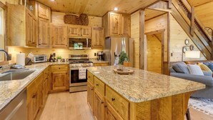 A wooden cabin kitchen with granite countertops, wooden cabinets, a central island, and a staircase leading to a loft.