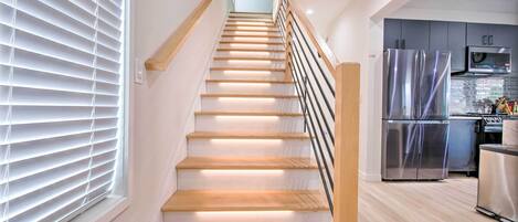 Un escalier avec des marches en bois et une rampe en métal, menant à un espace cuisine équipé d’appareils électroménagers en acier inoxydable.