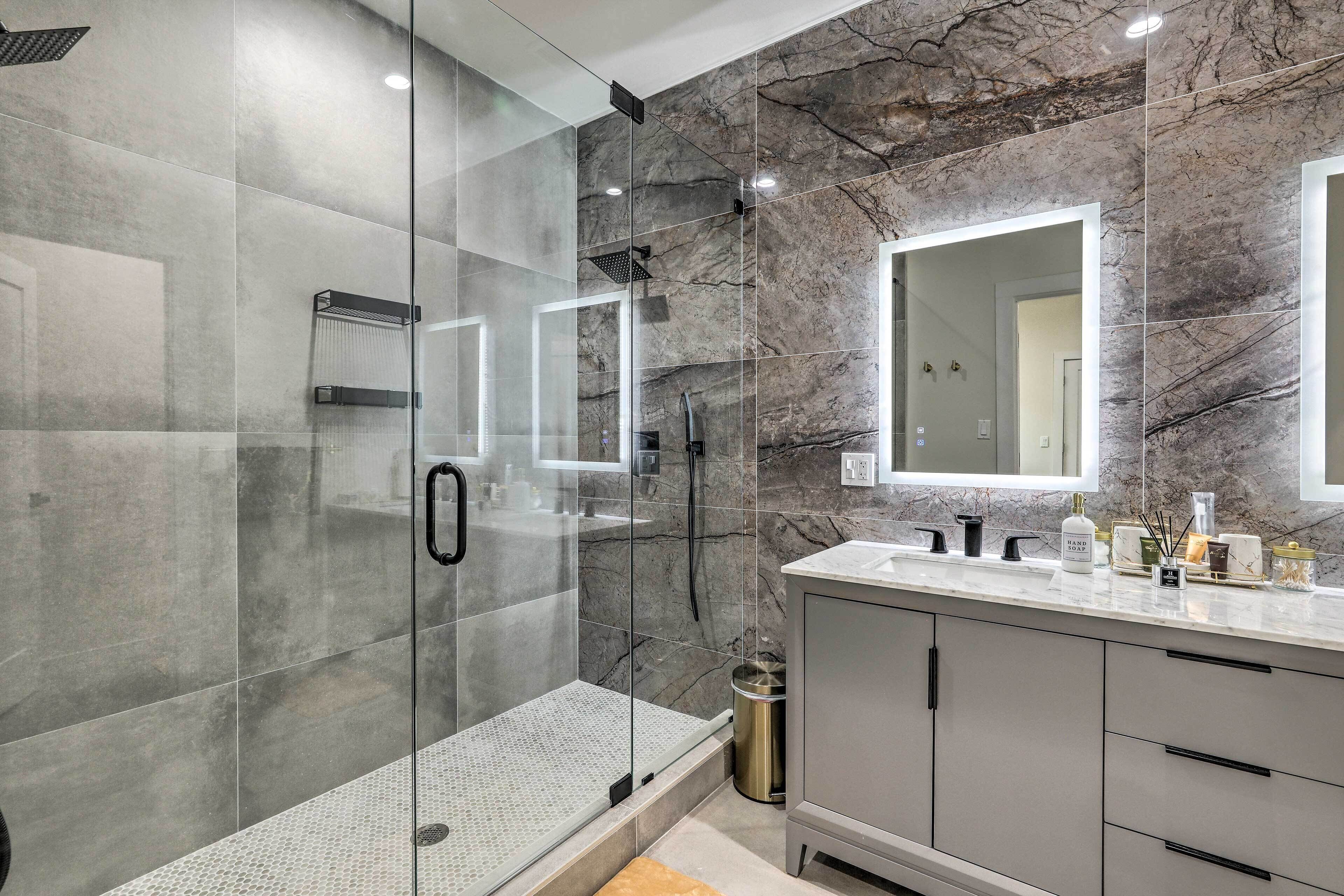 A modern bathroom with a glass-enclosed shower, marble walls, a large mirror, and a vanity with toiletries.
