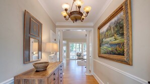 A hallway with a wooden dresser, a chandelier, framed artwork, and a large landscape painting. - Stunning Lake Oconee Retreat w/ Pool Access! (Eatonton)