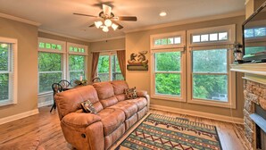 A living room with a leather sofa, a ceiling fan, large windows, a fireplace, and a dining area with a table and chairs. - Hiawassee Haven on Lake Chatuge w/ Boat Slip! (Hiawassee)