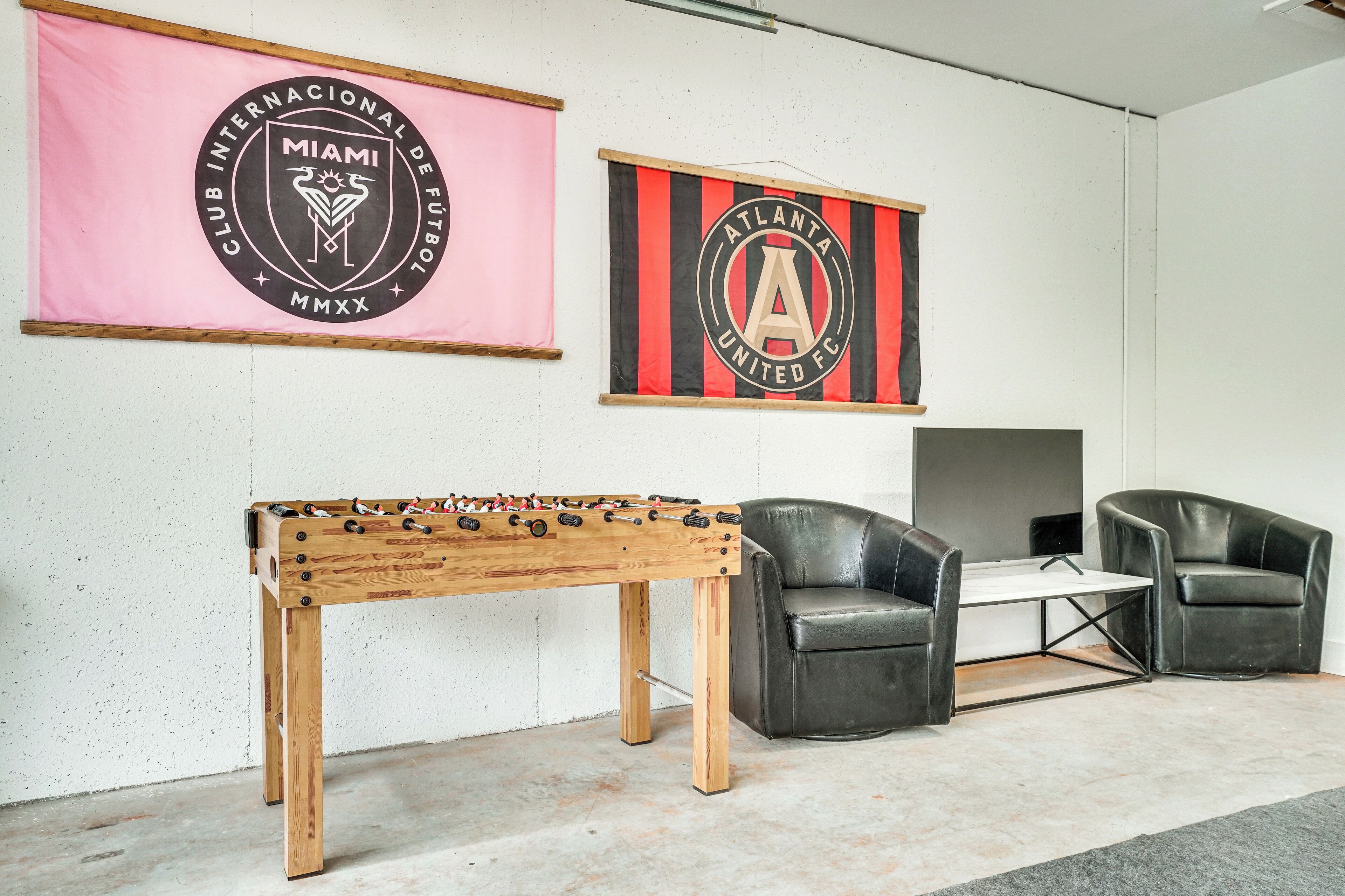 Una habitación con mesa de fútbol, dos sillones de cuero y dos banderas en la pared.