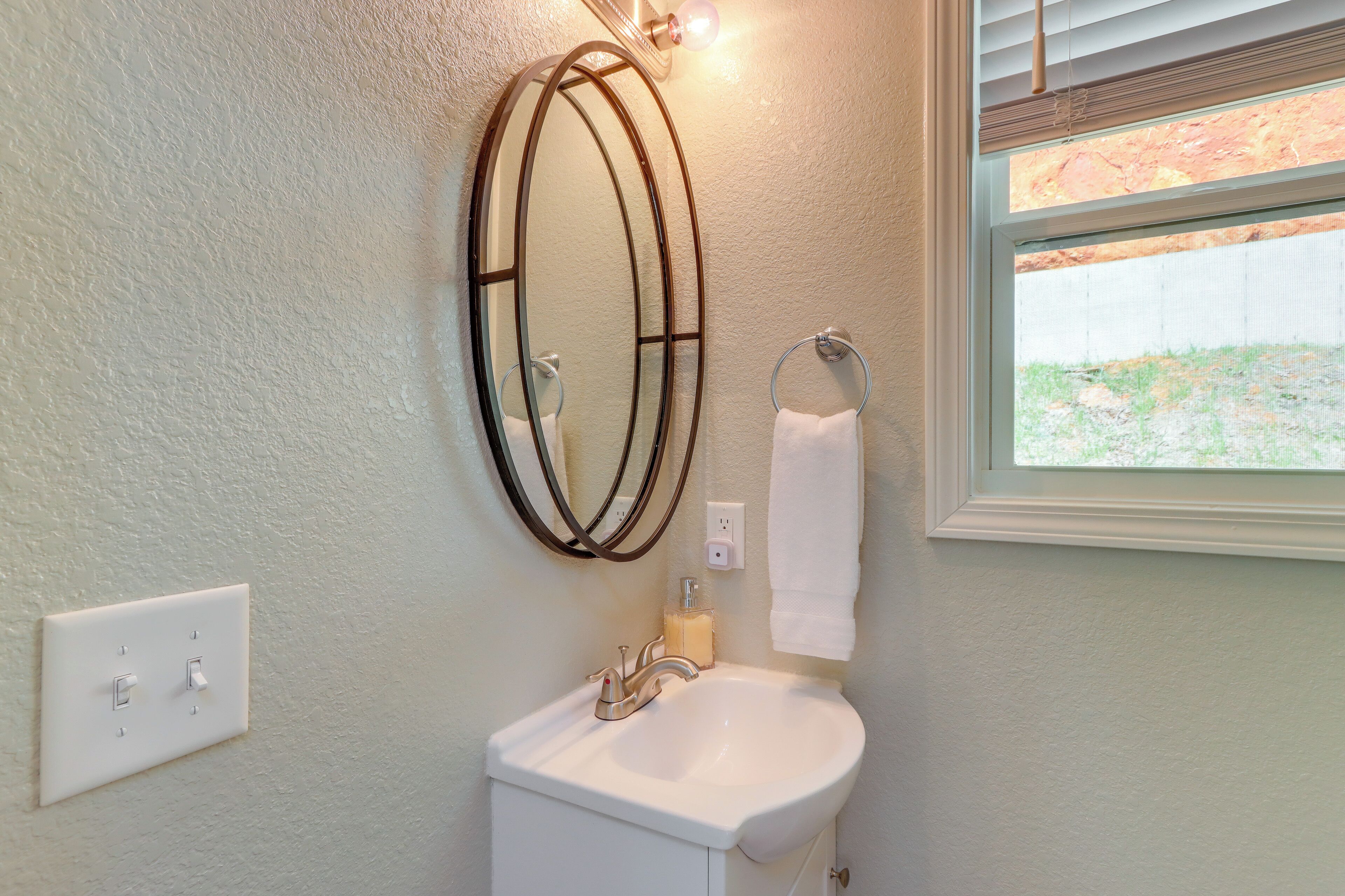 A bathroom with a white sink, a mirror with a metal frame, a towel hanging on a rack, and a window with blinds.