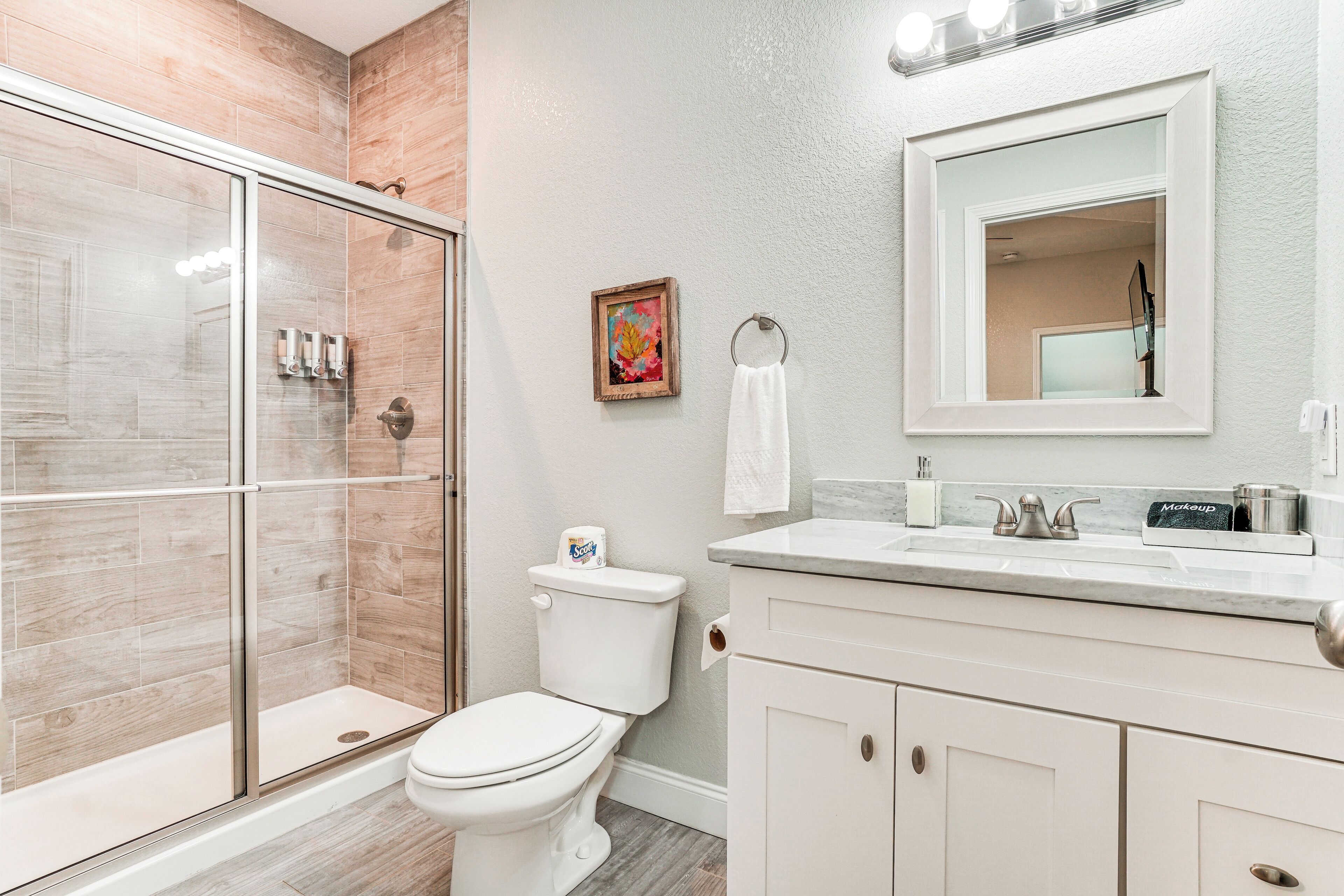 A bathroom with a glass shower enclosure, a toilet, a sink with a mirror, and a framed picture on the wall.