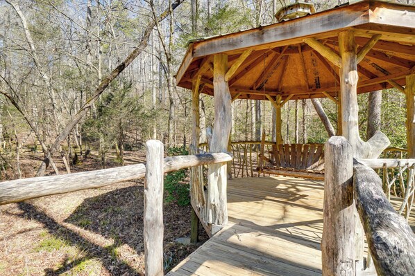 A wooden gazebo with a bench inside, surrounded by a wooden deck and a fence, set in a forested area.
