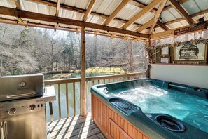 A covered outdoor deck with a hot tub, a grill, and a view of a forested area.